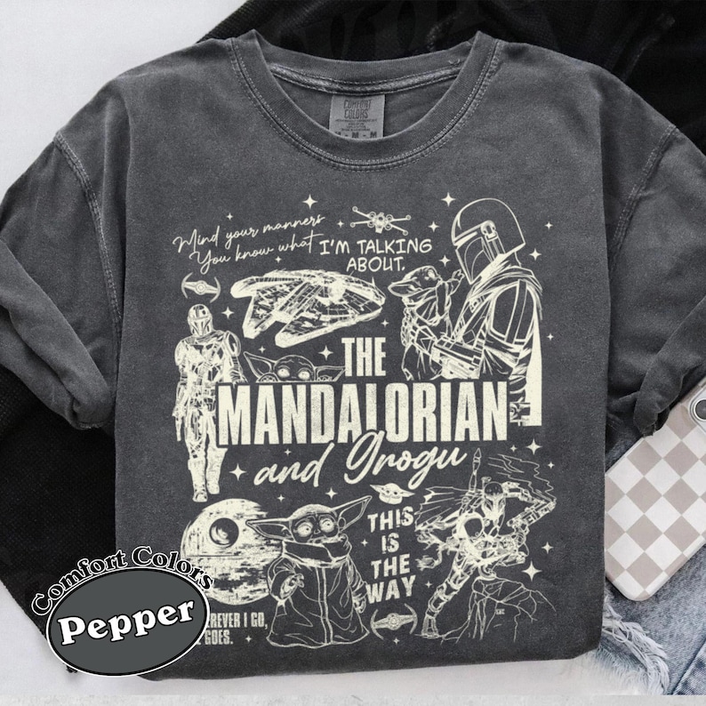 May include: Dark grey Comfort Colors t-shirt with a graphic design featuring characters and text from The Mandalorian series. The design includes the text "Mind your manners, You know what I'm talking about." and "The Mandalorian and Grogu."