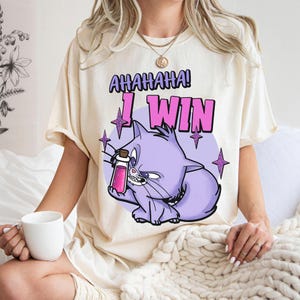 Puede incluir: Camiseta color crema con un gato de dibujos animados sosteniendo una botella de poción. El gato es morado, con el texto "AHAHAHA! I WIN" en letras de burbujas. Acentos de estrellas moradas.