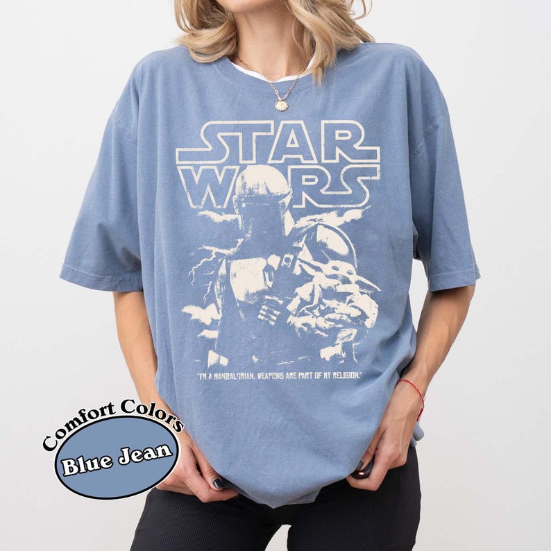 Puede incluir: Camiseta azul vaquero con un gr&aacute;fico de Star Wars. El dise&ntilde;o presenta el logotipo de Star Wars y una ilustraci&oacute;n de un personaje. El texto "I'M A MANDALORIAN, WEAPONS ARE PART OF MY RELIGION" est&aacute; impreso debajo del gr&aacute;fico.