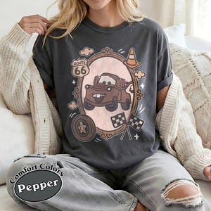 May include: Dark gray t-shirt featuring a cartoon image of a brown tow truck inside an ornate frame, with Route 66 signage and checkered flags. The shirt also has the words "Comfort Colors" and "Pepper" printed on it.