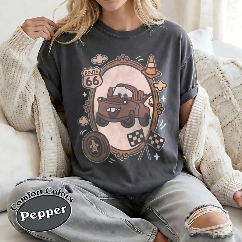 May include: Dark gray t-shirt featuring a cartoon image of a brown tow truck inside an ornate frame, with Route 66 signage and checkered flags. The shirt also has the words "Comfort Colors" and "Pepper" printed on it.