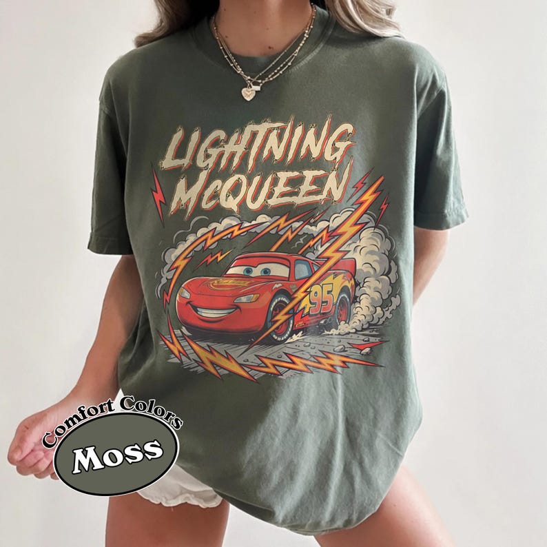 May include: Moss green t-shirt featuring a graphic of Lightning McQueen from the Cars movie. The design includes the text "Lightning McQueen" and the number "95" on the red race car. The shirt is a Comfort Colors brand.