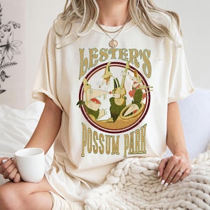 May include: Cream-colored t-shirt featuring a vintage-style graphic. The design includes the text "Lester's Possum Park" in a circular emblem with cartoon possums playing instruments. The shirt has short sleeves.
