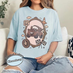 May include: Light blue t-shirt with a cartoon graphic of a brown truck inside a decorative frame, Route 66 sign, and checkered flags. The shirt has the words "Comfort Colors Chambray" printed on it.