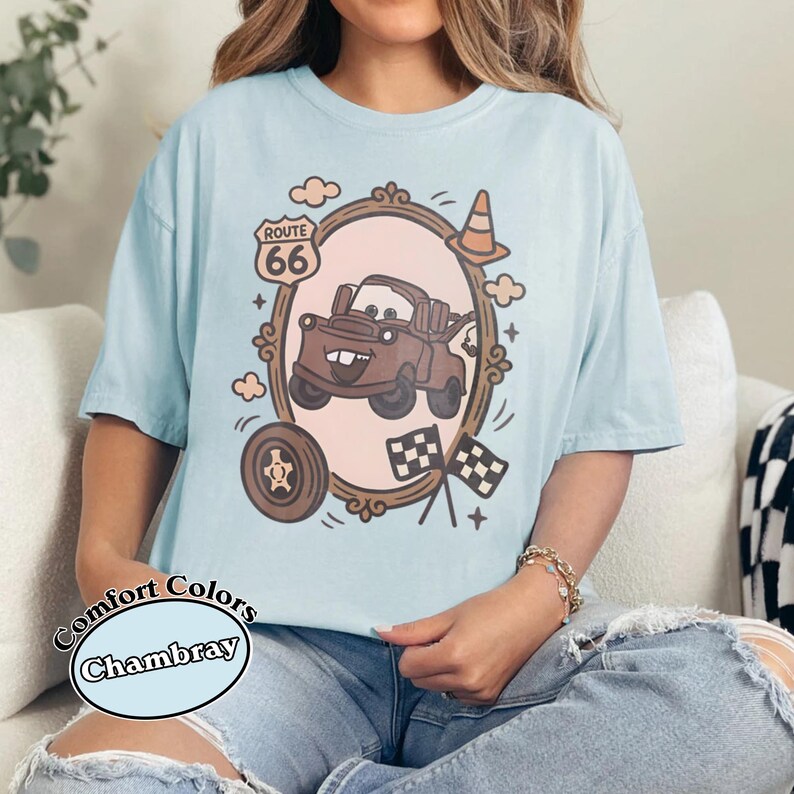 May include: Light blue t-shirt with a cartoon graphic of a brown truck inside a decorative frame, Route 66 sign, and checkered flags. The shirt has the words "Comfort Colors Chambray" printed on it.
