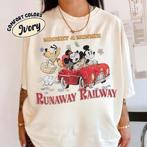 May include: Ivory-colored t-shirt featuring a graphic of Mickey and Minnie Mouse in a red car with Pluto. The text "Mickey & Minnie" and "Runaway Railway" are printed on the shirt. The shirt is labeled "Comfort Colors".