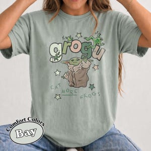 May include: Sage green t-shirt featuring a Grogu graphic with the text "Eat More Frogs." The design includes Grogu, stars, and frogs. The shirt is made of a soft, comfortable material.