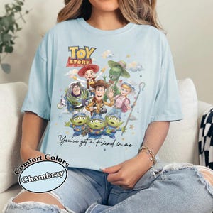 Pu&ograve; includere: T-shirt azzurra con una grafica di Toy Story con Woody, Buzz Lightyear e i piccoli omini verdi. Il testo "Toy Story" &egrave; in alto, con "You've got a Friend in me" sotto. La maglietta &egrave; etichettata "Comfort Colors Chambray".