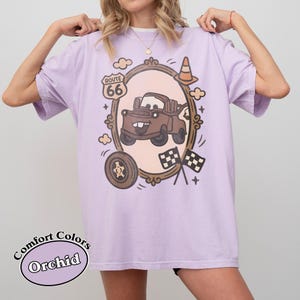 May include: Orchid-colored t-shirt featuring a cartoon image of a brown tow truck inside an ornate frame, with Route 66 signage, a traffic cone, and checkered flags. The shirt also has the words "Comfort Colors" and "Orchid" printed on it.