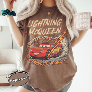 May include: Espresso-colored t-shirt featuring Lightning McQueen from the Cars movie. The graphic shows the red race car with the number 95, surrounded by lightning bolts and smoke. The shirt also has the words "Lightning McQueen" in a distressed font.