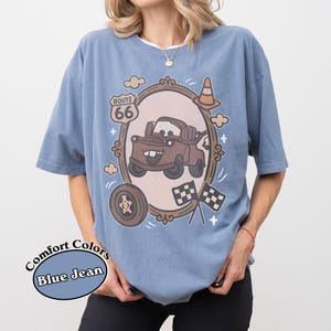May include: Blue jean Comfort Colors t-shirt featuring a cartoon image of Tow Mater from the movie Cars. The design includes a Route 66 sign, a tire, checkered flags, and a traffic cone. The shirt has short sleeves and a crew neck.