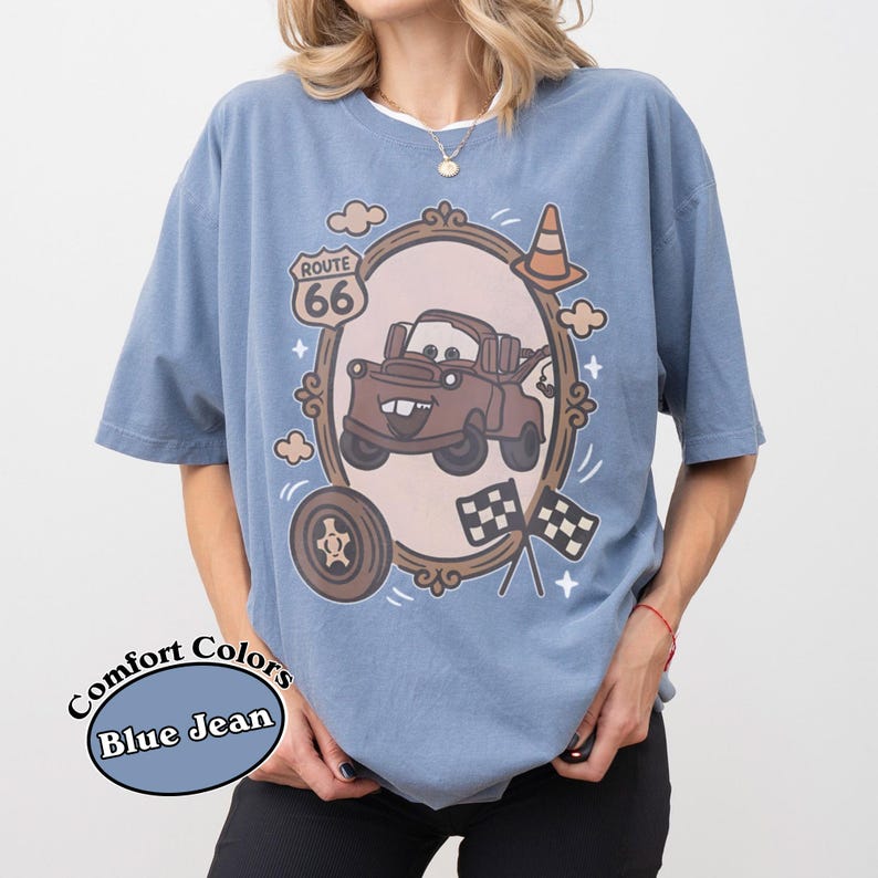 May include: Blue jean Comfort Colors t-shirt featuring a cartoon image of Tow Mater from the movie Cars. The design includes a Route 66 sign, a tire, checkered flags, and a traffic cone. The shirt has short sleeves and a crew neck.
