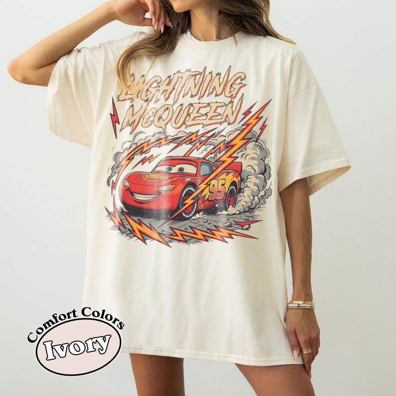 May include: Ivory-colored t-shirt featuring a graphic of Lightning McQueen from the movie Cars. The design includes the text "Lightning McQueen" and the number "95" on the car. The shirt is a Comfort Colors brand.