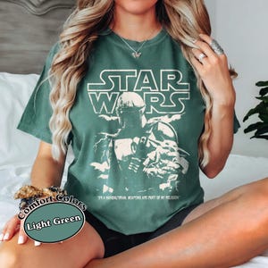 Puede incluir: Camiseta verde claro con un gr&aacute;fico desgastado de Star Wars que presenta una imagen de un Mandaloriano. El texto "STAR WARS" est&aacute; encima de la imagen, y la frase "I'M A MANDALORIAN, WEAPONS ARE PART OF MY RELIGION" est&aacute; debajo. La camiseta est&aacute; etiquetada como "Comfort Colors" y "Light Green".