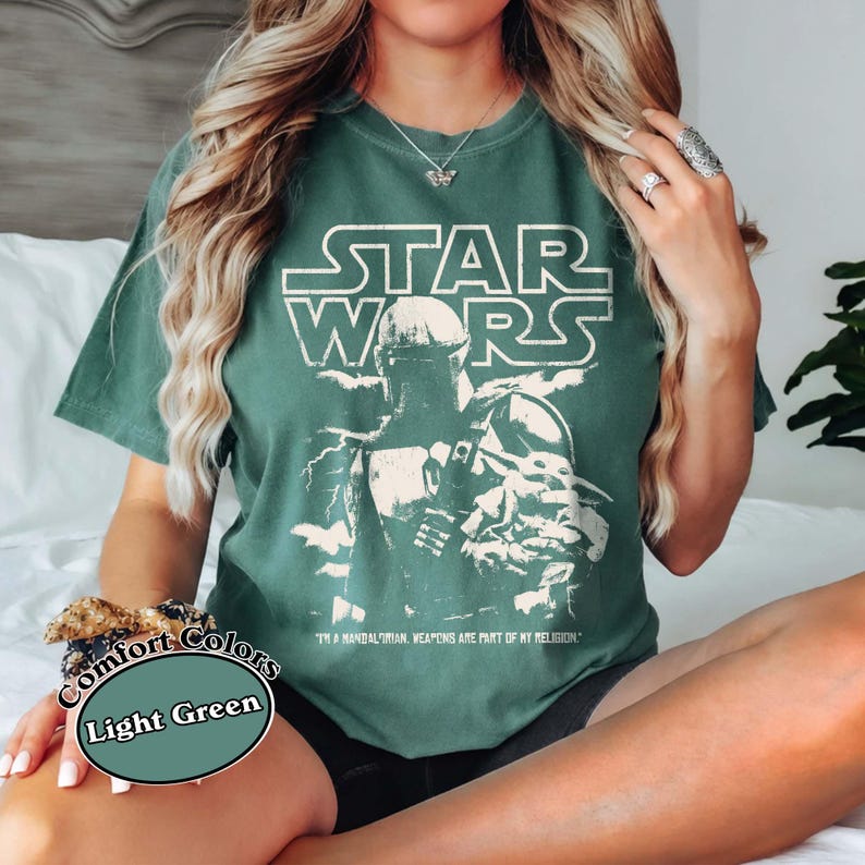 Puede incluir: Camiseta verde claro con un gr&aacute;fico desgastado de Star Wars que presenta una imagen de un Mandaloriano. El texto "STAR WARS" est&aacute; encima de la imagen, y la frase "I'M A MANDALORIAN, WEAPONS ARE PART OF MY RELIGION" est&aacute; debajo. La camiseta est&aacute; etiquetada como "Comfort Colors" y "Light Green".