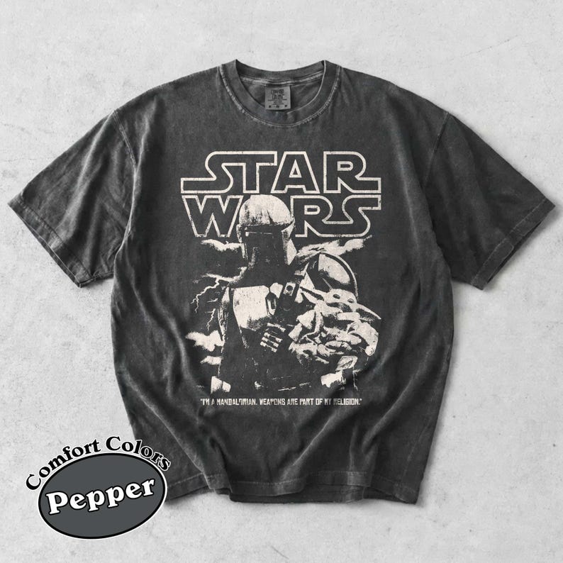 Puede incluir: Camiseta gris oscuro con un gr&aacute;fico de The Mandalorian y Grogu de Star Wars. El logotipo de Star Wars est&aacute; encima de la imagen, con texto debajo. La camiseta es una camiseta Comfort Colors Pepper.