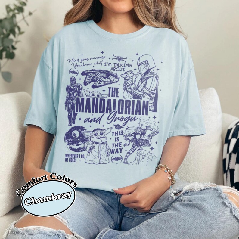 May include: Pale blue t-shirt with illustrations of characters from "The Mandalorian" series. The shirt features the text "The Mandalorian and Grogu" and other phrases. The shirt is a Comfort Colors Chambray.