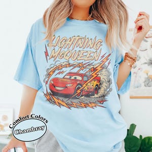 May include: Light blue Comfort Colors Chambray t-shirt featuring a graphic of Lightning McQueen from the Cars movie. The design includes the text "Lightning McQueen" and the number "95" on the red race car, with lightning bolts and smoke effects.
