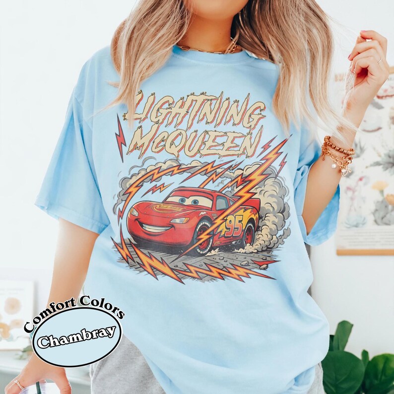 May include: Light blue Comfort Colors Chambray t-shirt featuring a graphic of Lightning McQueen from the Cars movie. The design includes the text "Lightning McQueen" and the number "95" on the red race car, with lightning bolts and smoke effects.