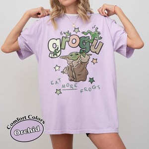 May include: Orchid-colored t-shirt featuring a Grogu graphic with the text "Eat More Frogs." The design includes Grogu, frogs, and stars. The shirt is a Comfort Colors brand.