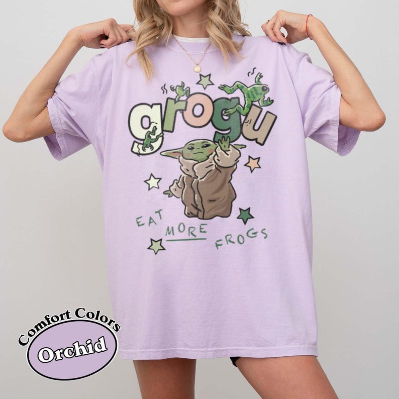May include: Orchid-colored t-shirt featuring a Grogu graphic with the text "Eat More Frogs." The design includes Grogu, frogs, and stars. The shirt is a Comfort Colors brand.