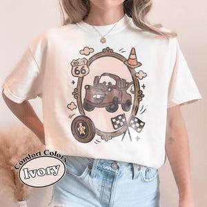May include: Ivory Comfort Colors t-shirt featuring a brown cartoon car design inside a decorative frame with Route 66 signage. The design includes a tire, clouds, and checkered flags. The shirt also has the words "Comfort Colors Ivory" printed on it.