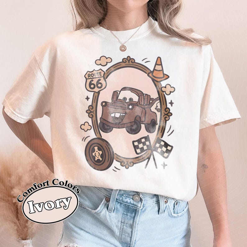 May include: Ivory Comfort Colors t-shirt featuring a brown cartoon car design inside a decorative frame with Route 66 signage. The design includes a tire, clouds, and checkered flags. The shirt also has the words "Comfort Colors Ivory" printed on it.