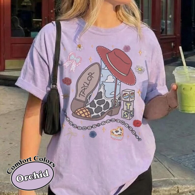 Puede incluir: Una camiseta de manga corta de color morado claro con un estampado gr&aacute;fico de una bota de vaquero con patr&oacute;n de vaca, un sombrero de vaquero rojo, el nombre Taylor, el n&uacute;mero 13 y bloques apilados con las letras T, S y S.