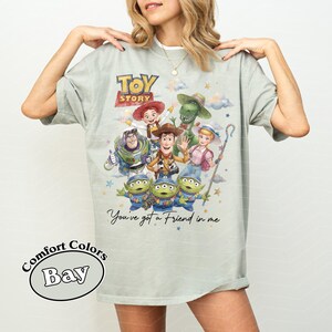 Pu&ograve; includere: T-shirt verde chiaro con una grafica colorata dei personaggi del film "Toy Story". Il design include Woody, Buzz Lightyear e altri personaggi, con il testo "Toy Story" e "You've got a Friend in me".