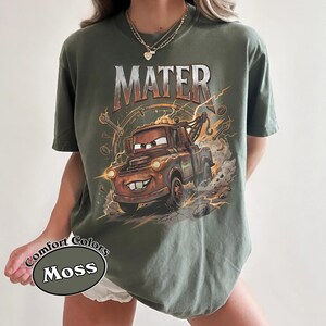 May include: Moss green t-shirt featuring a graphic of Mater from the movie Cars. The design includes the word "MATER" in a distressed font above the cartoon tow truck. The shirt is a Comfort Colors brand.