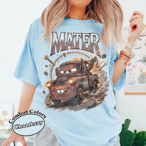 May include: Light blue t-shirt featuring a graphic of Mater from the movie Cars. The design includes the word "MATER" in a distressed font above the character. The shirt is labeled "Comfort Colors Chambray".