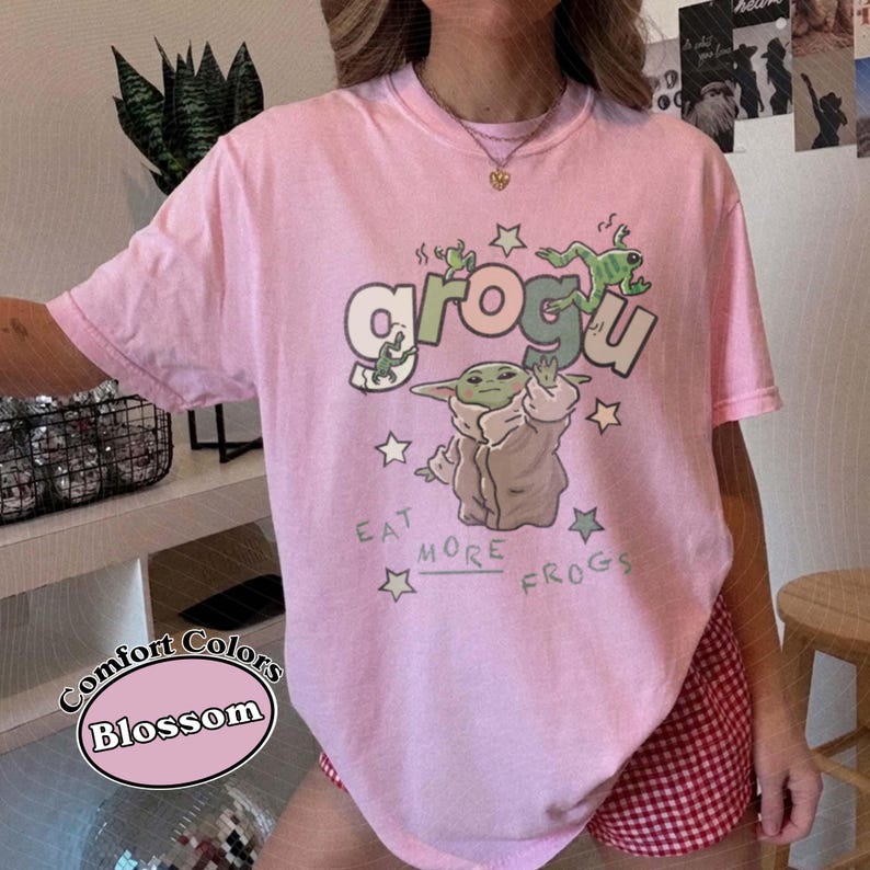 May include: A light pink t-shirt with the word "Grogu" in green and white, featuring a cartoon character and the phrase "Eat More Frogs." The shirt is a Comfort Colors Blossom tee.