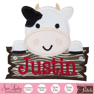 May include: Embroidered iron-on patch featuring a cartoon cow peeking over a wooden sign with the name "Justin" in red lettering. The sign has a brown and white striped pattern.
