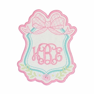 May include: Embroidered patch in a shield shape with a pink bow at the top and a monogram in the center. The patch has a white background with a light blue border and floral accents.