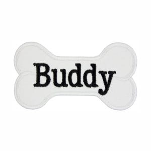 May include: A white bone-shaped embroidered patch with a stitched border. The word Buddy is centered on the patch in black, bold, serif lettering. The fabric has a visible woven texture throughout the white surface of the bone shape.