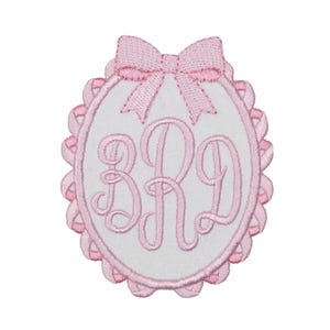 May include: An oval-shaped embroidered patch featuring a white center with the pink monogram letters BRD in a cursive script. The patch is bordered by a scalloped pink edge and topped with a pink embroidered bow detail at the very top.