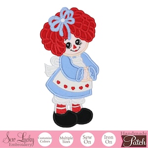 May include: A handmade patch featuring a Raggedy Ann doll design. The doll has red yarn hair, a blue bow, and a blue dress with a white apron adorned with red hearts. The patch has black shoes and white socks with red stripes.