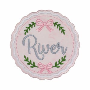 May include: A circular embroidered patch with a scalloped edge, featuring a light pink background. The name River is stitched in grey cursive script in the center, surrounded by two pink bows and green leafy branches at the top and bottom.