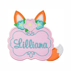 May include: A pink embroidered patch in the shape of a fox with the name "Lilliana" in teal script. The fox has orange ears, a white inner ear, and a fluffy orange and white tail. The patch is decorated with teal and pink flowers.