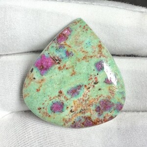 Puede incluir: Una piedra preciosa pulida con forma de lágrima. La piedra presenta un color base verde claro con parches de rosa vibrante y turquesa. Las motas marrones y negras añaden detalles a la superficie.