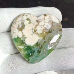 May include: Heart-shaped gemstone with a unique pattern. The stone features a mix of colors, including white, green, and brown. The white areas have a cloud-like appearance, while the green and brown sections create a natural, earthy look. The polished surface reflects light.