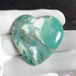 Può includere: Una pietra a forma di cuore con una superficie lucida. La pietra presenta una miscela di colori, tra cui tonalità di verde, bianco e grigio. Il cuore è tenuto in un guanto bianco, evidenziando la sua forma e le variazioni di colore.