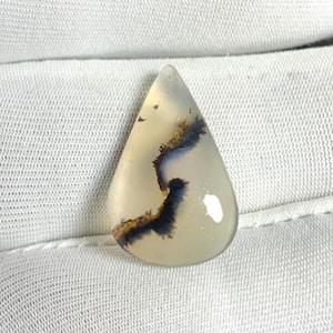 Può includere: Una gemma a forma di goccia con una base bianco crema traslucida. La pietra presenta intricate inclusioni dendritiche marrone scuro e nere che assomigliano a un paesaggio naturale. La superficie lucida riflette la luce.