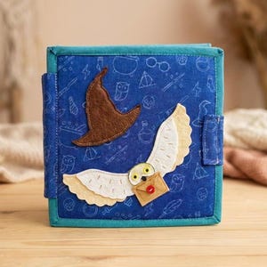 May include: A blue fabric book with a whimsical design. The cover features a brown sorting hat and a white owl with a letter. The background is patterned with magical symbols. The book is trimmed in teal.