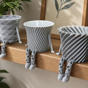 May include: Three gray decorative planters with attached legs, sitting on a wooden shelf. The planters have unique geometric and spiral designs. A framed picture and potted plants are in the background.