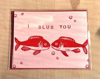 May include: A rectangular greeting card featuring a light pink background with two red fish facing each other. The text I BLUB YOU is printed in red above the fish, accompanied by small red circular bubbles scattered across the card surface.