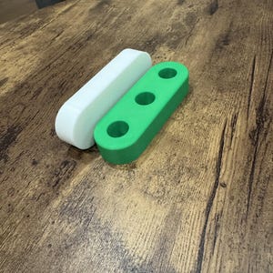 May include: Two elongated, rounded rectangular blocks are positioned side by side. One block is solid white, while the other is bright green and features three evenly spaced circular holes drilled through its top surface along the center line.