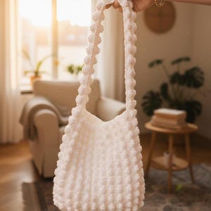 以下が含まれることがあります： 白いかぎ針編みのトートバッグ。テクスチャード加工のバブルステッチのデザイン。バッグには、一連のバブルが付いた2つの長いハンドルが付いています。バッグは、太陽光が差し込む窓の前に掲げられています。