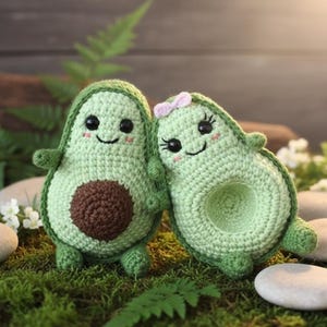 Puede incluir: Dos peluches de aguacate de ganchillo, uno con un lazo rosa, tomados de la mano. Los aguacates son de color verde claro con centros marrones y caras sonrientes. Se colocan sobre un lecho de musgo verde con piedras blancas y vegetación.