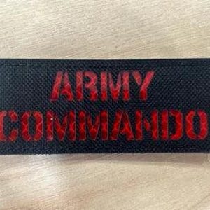 May include: A rectangular black patch with the words "ARMY COMMANDO" in red, textured lettering. The patch is on a light brown, wood-grain surface.